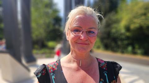 Claire Sharpe smiles at camera, she is wearing a black floral dress and a pair of glasses. The structure is behind her.