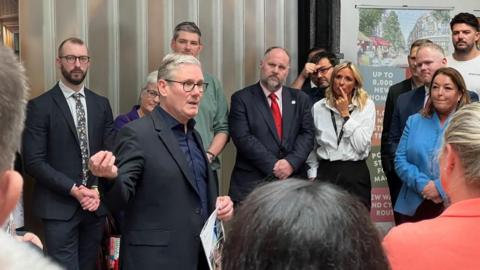 Prime Minister Sir Keir Starmer, wearing a black jacket and dark blue shirt, gives a speech to small business owners and supporters in Swindon. Thirteen people can be seen watching on.