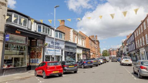Several cars parked on Grange Road. Shops run along both sides of the street which has old terrace buildings of two and three storeys. There is yellow bunting stretching above the road.