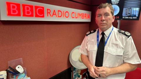 Library image of Rob Carden, chief constable of Cumbria Police, pictured in one of BBC Radio Cumbria's studios. He is wearing his police uniform - a white shirt with black tie. On the wall to his right-hand side is a large BBC Radio Cumbria sign.