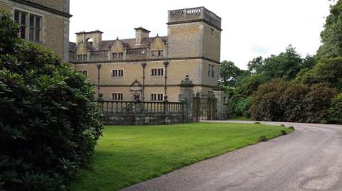 A large, grey, three-storey stone brick building with a large rectagonal tower in the corner and a curving drive leading to a gateway through a stone fence