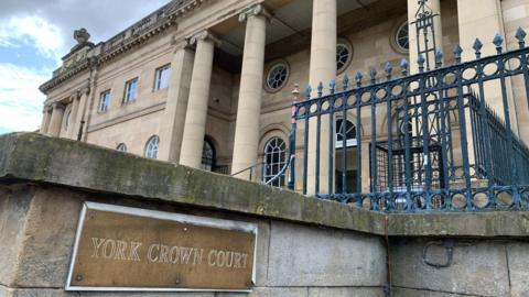 The outside of a court building, with a small sign reading: "York Crown Court." A blue metal fence runs around the side of the grand building, which has circular, oval and square windows. 