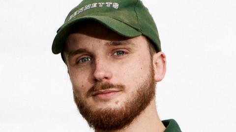 Stephen Bennett, a young man with a reddish beard, is pictured looking at the camera. He is wearing a green cap with "BENNETTS" printed on it.
