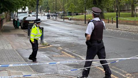 Two police officers stand by the side of a section of the road which has been cordoned off