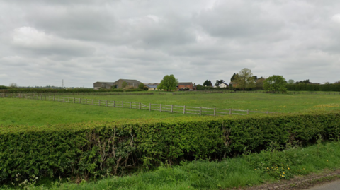 A green hedgerow with green fields behind and farm buildings in the background