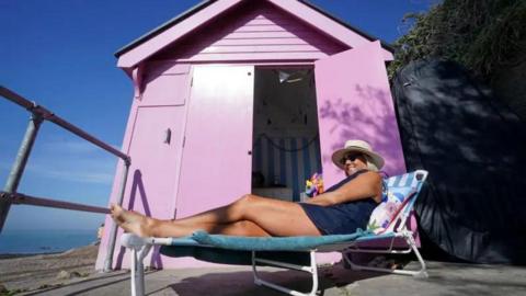 A woman sat on a sun lounger outside a pink beach hut by the seafront in  Folkestone.