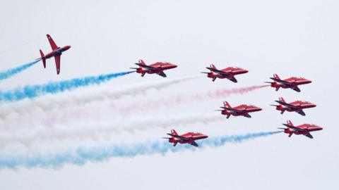 The Red Arrows at an air display. The red aircraft are trailing red, white and blue smoke