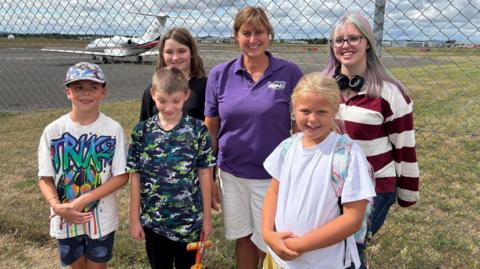 A group of five children, aged between 10 and 17, standing near a runaway at Gloucestershire Airport. There are two young boys on the left and three older girls on the right. In the middle is a woman from Gloucestershire Young Carers who is wearing a purple polo shirt and white shorts.