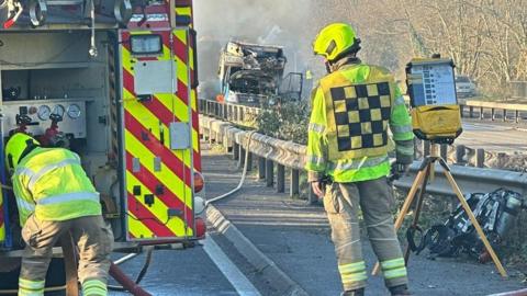 Backs of two firefighters standing on a road with a fire appliance a burnt, smoking van can be seen in the distance.