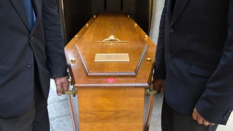 A generic image of two men in black suits, photographed from the chest down, holding a wooden coffin between them