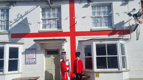 Man in a knight outfit with a cape and chainmail next to a woman dressed like a London beefeater. Behind them is a white building which two red stripes which looks like an England flag