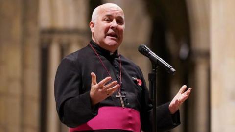 Archbishop of York standing and holding his hands open, palms upwards, wearing his black cassock with red trim