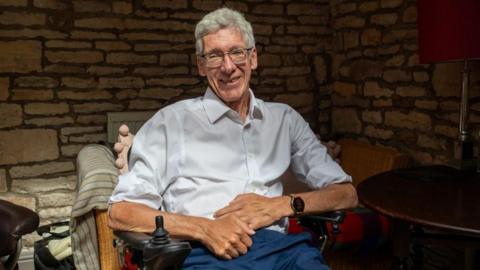 Councillor Tony Dale smiles at the camera. He sits in a chair while wearing a white shirt and blue trousers. His sleeves are rolled up to just above his elbows. He wears rectangular glasses and a watch on his left wrist. A brick wall can be seen behind him. 