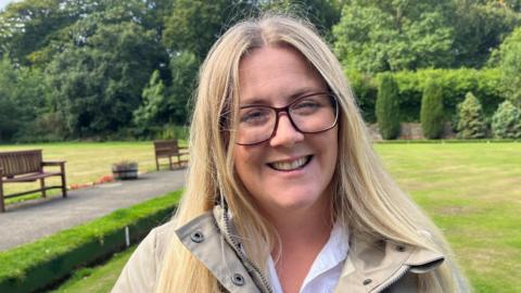A woman with long blonde hair, wearing glasses and a cream coat smiles into the camera, she is stood in a park with benches and hedgerows behind her.