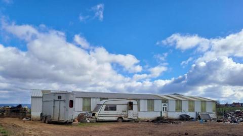 A wide steel frame building with white and green panels. A caravan is sat in front. Blue skies and clouds can be seen in the background.