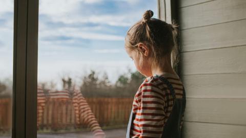 Little girl with bunches looking out a large window, inside a green wooden hut. 