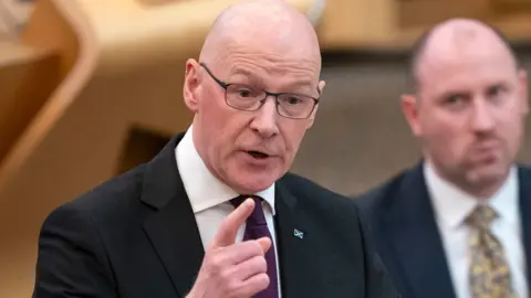 First Minister John Swinney gesticulates with a finger while wearing a suit and tie. In the background, sitting, is Health Secretary Neil Gray.