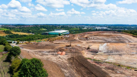 Image shows a large swathe of land that has been taken back to soil level which is surrounded by trees and fields. There are tyre and caterpillar tracks crisscrossing the land, which looks like the beginning of a building site.