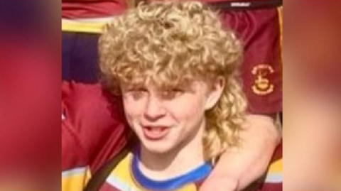 Harry, a young boy with curly blonde hair, wearing a red and yellow top. Someone has their arm around him. It is a close-up from a group photo, and there are other people in the team kit around him.