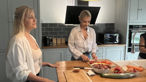 Two blonde women prepare food on a wooden surface in a kitchen