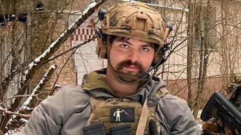 A person wearing tactical gear and a helmet stands outdoors in a snowy, wooded area, holding a rifle in front of an abandoned-looking building.