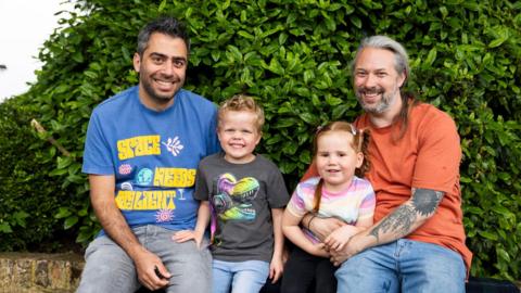 Luke and Alex sitting on a brick wall with a hedge behind them. In the middle are their two young daughters. They are all smiling at the camera.