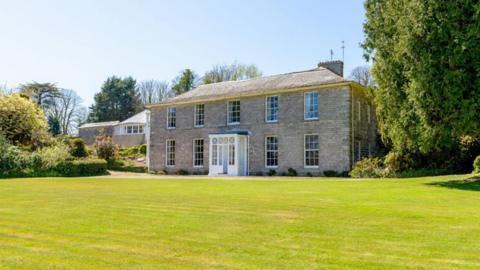The picture shows a large Georgian country house built from pale stone, set back behind a wide, neatly cut green lawn. The house is symmetrical and two storeys high, with a central white front door and evenly spaced sash windows on either side and above. The roof is shallow and slate‑coloured, with chimneys visible at either end. In the foreground, the lawn stretches right up to the front of the building