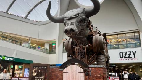 A photo of a mechanical bull in the middle of a train station, it has a flat cap on and is grey, matching the colour of it's head. 