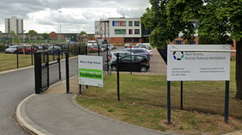 The exterior of Blacon High School shows two signs and a staff car park. In the background there is a brick two-storey building and to the left and right there are grass fields.