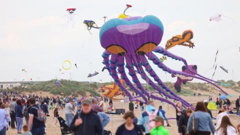 A large kite in the shape of an octopus being flown on Redcar beach. It is purple with large orange eyes. A number of smaller kites are also being flown in the area with dozens of people looking on.