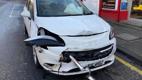 A white car which has dents all over the bonnet and is missing a front light.