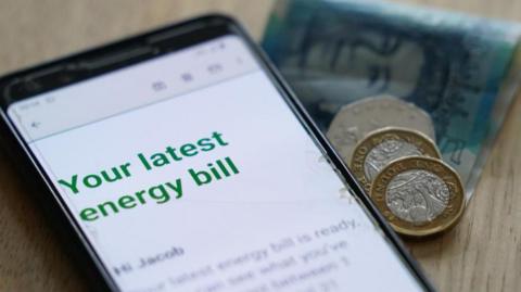 A phone with an email reading "your latest energy bill" on the screen, sat on a wooden table next to a greeny-blue five pound note, two pound coins and a 50p coin.