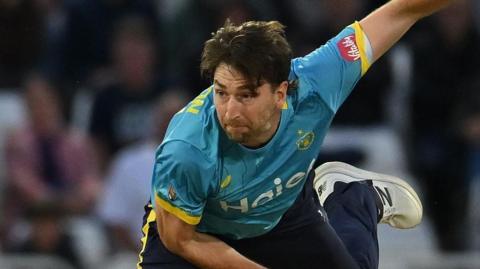 Bowler Richard Gleeson, wearing a blue shirt and navy trousers, watches after bowling a ball while playing cricket for Warwickshire Bears