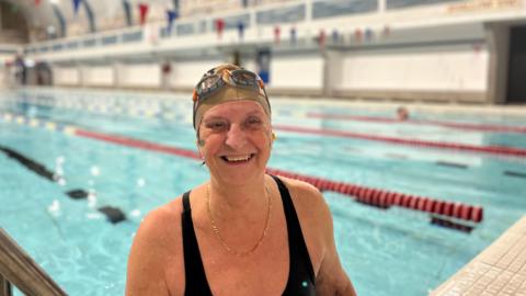 A swimmer stands in the pool, smiling at the camera. She wears a swimming cap, googles, black swimsuit and gold necklace.