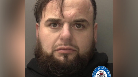 A mugshot of a man with a thick brown beard and brown hair. A grey background is behind him.