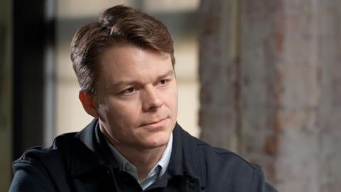 Palantir UK boss Louis Mosley speaking to the BBC. He is wearing a dark jacket with a button up jumper and is sitting in front of a grey wall.