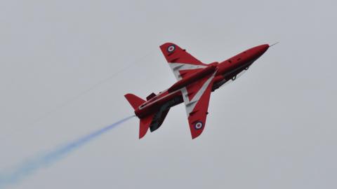 A red arrow plane soaring through grey skies with blue smoke trailing behind it.