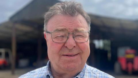 John Hardy looking to the camera, wearing glasses and a blue checked shirt. Behind him is a barn with tractors in