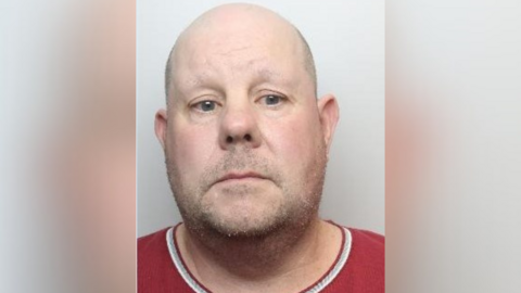 Mugshot of Sean Gamble wearing a red t-shirt. The collarless edge has a white line around it. He has a shaved head and stubble.  