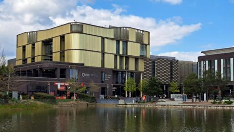 A large gold and black building that is several stories high and has large windows. There is a large pond not far from the building.