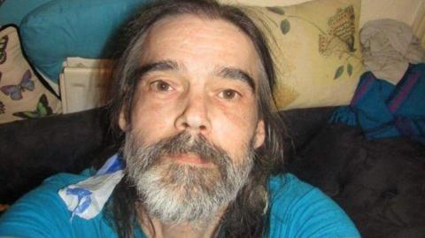 The photo shows a person taking a selfie from a high angle while sitting indoors. They are wearing a bright blue top. They have long dark hair and a greying beard. A piece of fabric is draped over one shoulder. Behind them is a dark-coloured sofa with cushions. One cushion has a butterfly print.