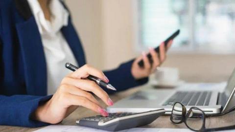Close up image of a professional woman sitting at a computer making calculations and looking at her smartphone. She is wearing a blue blazer and an open neck white blouse. (Stock image)