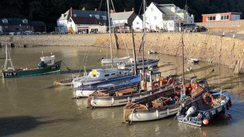 Nine boats of various use sit dotted about Minehead Harbour at high tide. They are penned in by a harbour wall which runs left to right of the shot. On the harbourside is a number of buildings - on the left is houses, and moving right there are more properties, a shop, a pub and a static caravan.