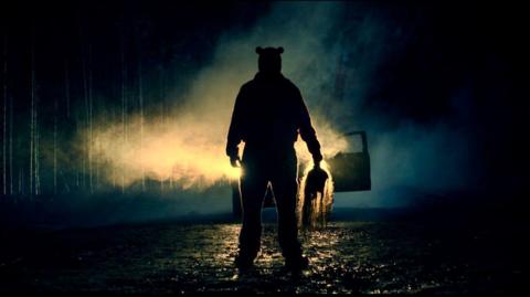 A silhouette of a figure in front of car headlights, at night. He is holding something ominous in his hand. 