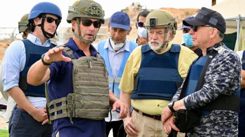 Wearing bullet proof vests and protective helmets, Mike Huckabee is flanked by officials gesturing to something beyond the camera