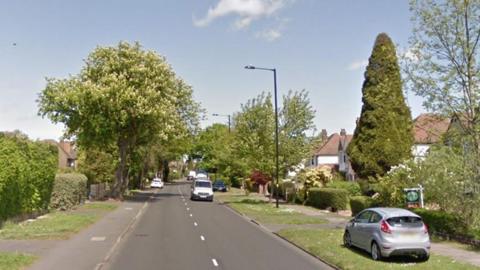 Hollyfield Road in Sutton Coldfield, a residential area with houses on both sides of the road, wide pavements and grass verges. There are parked cars and lampposts and several trees.