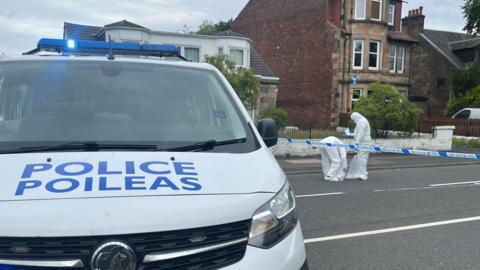 A police van and police tape on a road, with two people in forensics outfits behind the tape working away. 