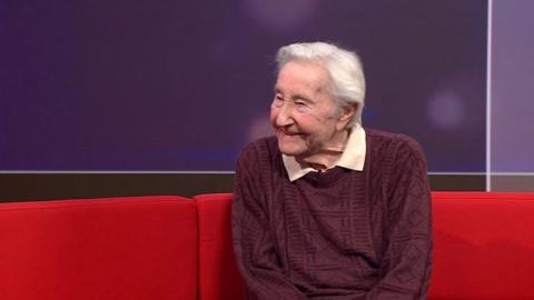 Edwin Rayner sits on a red sofa in the ˿ South studio, there is a purple background. He is smiling, looking away from the camera, has white hair in a side parting and wears a mauve cable knit jumper with a nude coloured polo top underneath.