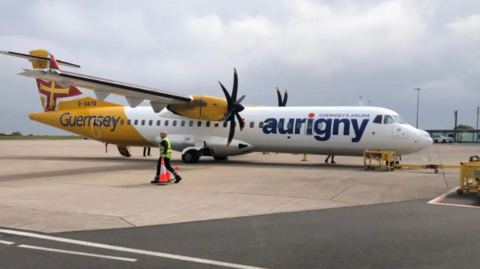 A yellow and white Aurigny plane on a runway in Guernsey with a person wearing high vis carrying traffic cones walking beside the plane on an overcast day