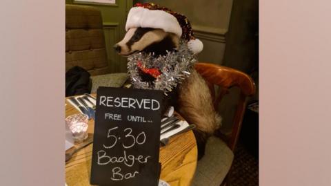 The stuffed badger Bertie wearing a Christmas hat and some tinsel around its head. It is at a pub table with a sign saying reserved for the Badger Bar staff party.